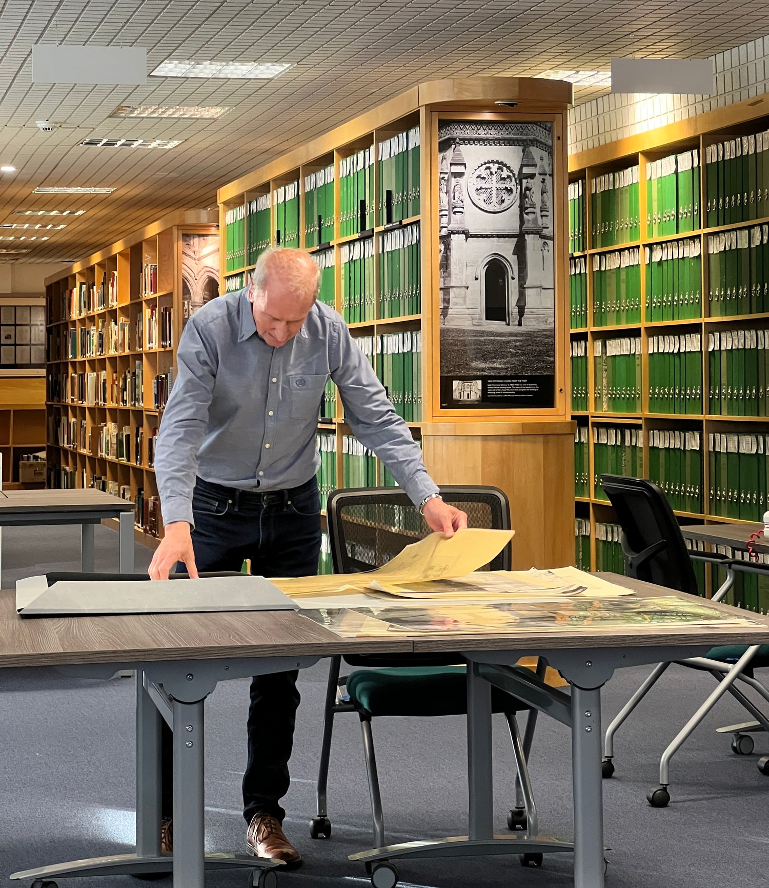A person stands over a table organising archival documents. In the background the room is filled with rows and shelves of green binders.